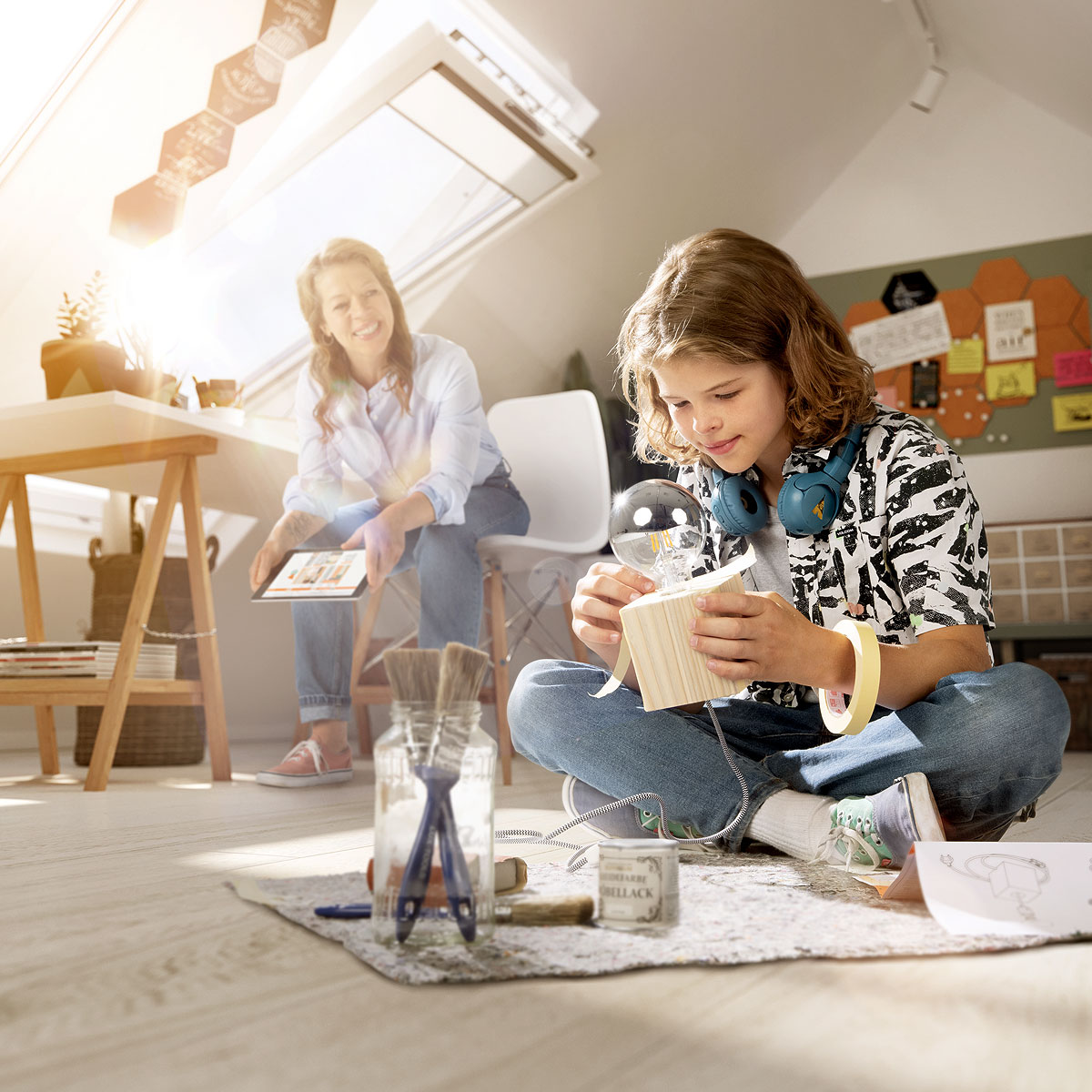 nju CGI, ein Junge sitzt auf dem Boden und bastelt eine DIY-Lampe zusammen, die Mutter sitzt im Hintergrund am Schreibtisch